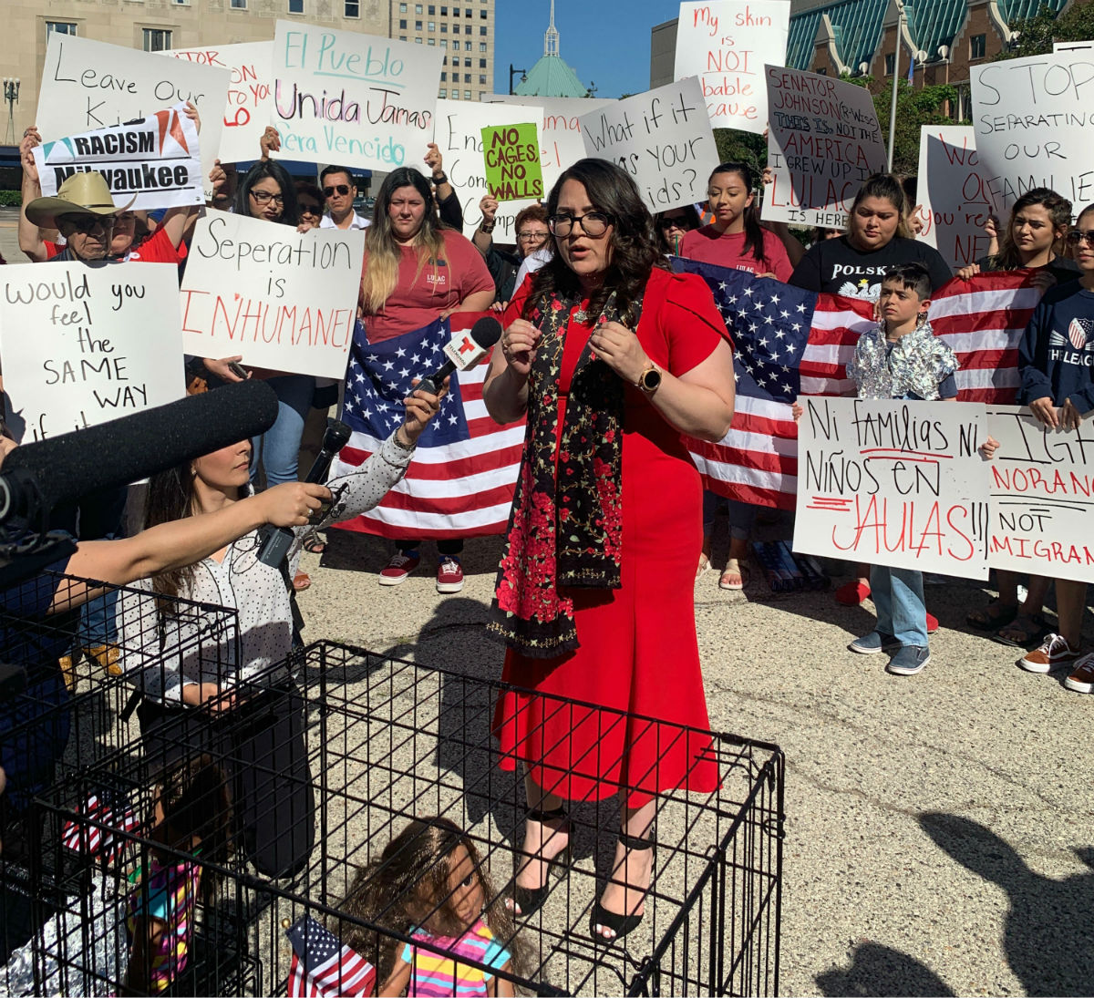 Protesters hold signs around baby dolls in cages, while one protester gives a speech