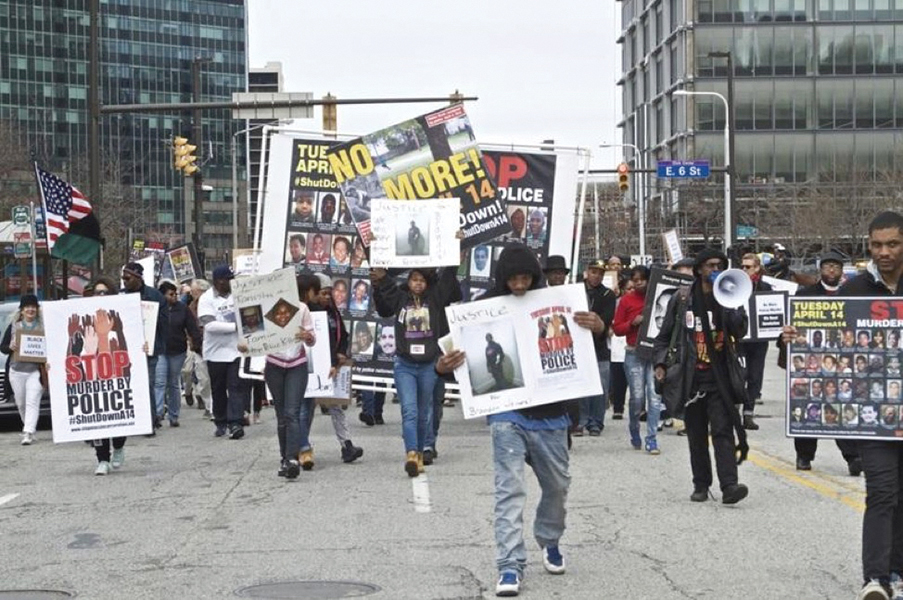 Photograph of people walking in a march protesting police violence in downtown Cleveland. Many people are carrying signs and one carries a megaphone. One sign reads, “Stop murder by police #ShutdownA14.”