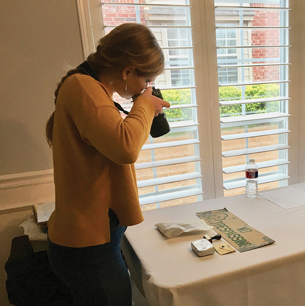 A young woman is standing near a window taking pictures of Girl Scout camp artifacts using a 35mm camera.