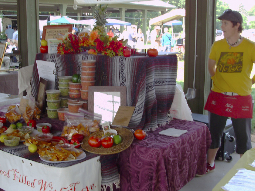 Stacks of salsa and tropical fruit on display. Samples of the salsa sit in front. A woman stands behind the display.