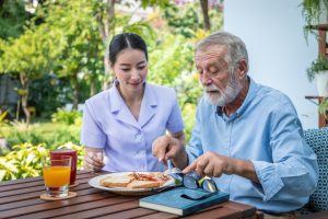 A health professional sits with an older man who is eating breakfast.