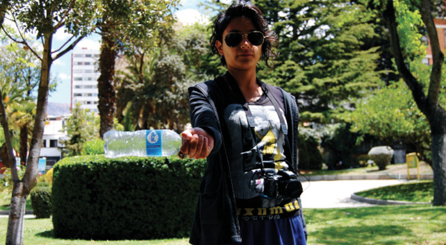 A young Latina artist wearing sunglasses and a camera around her neck is pictured outside with a plastic water bottle in hand, facing the camera as if to show the water bottle off; she stands in front of a verdant background.