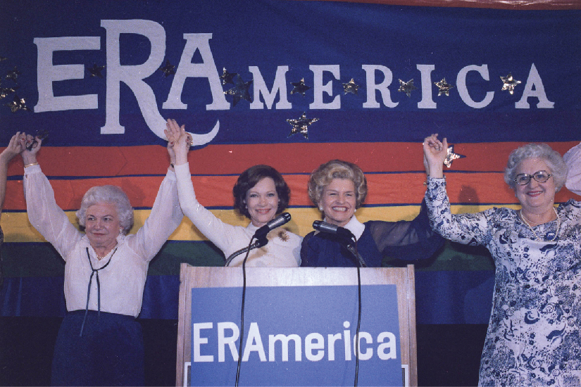 A photo of Rosalynn Carter and Betty Ford speaking at a rally in favor of the Equal Rights Amendment. Signs on stage read “ERAmerica.”