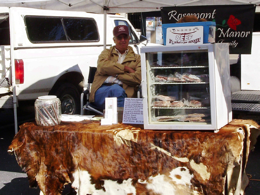 A man with his arms crossed, sits behind a cow skin covered stall. On the stall is a cooler filled with packed meats and a sign that reads, “Longhorn Beef Stall – It’s what’s for dinner!”