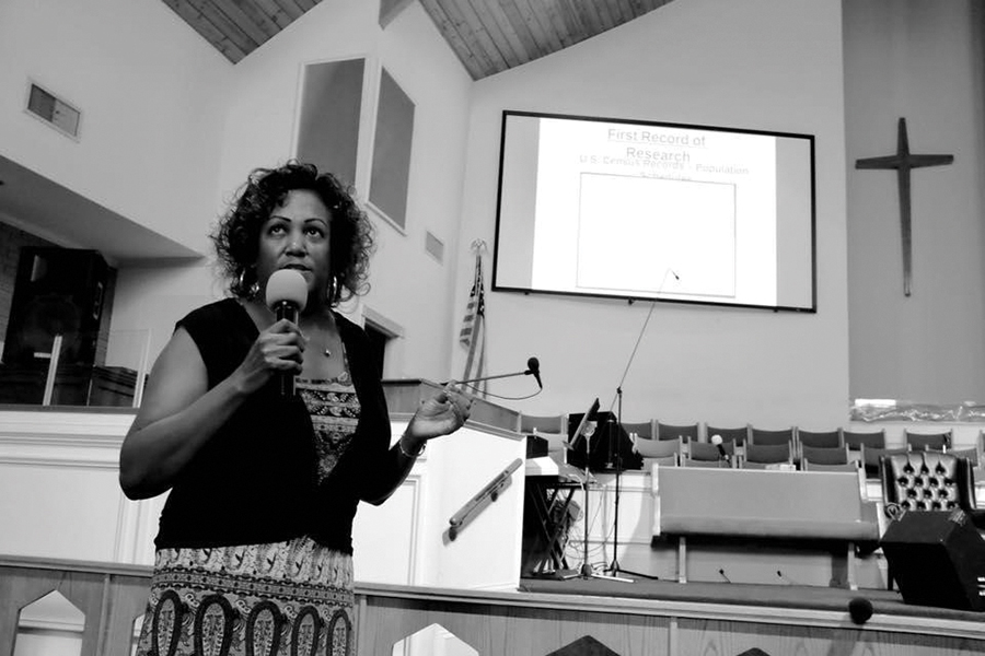 Hill is standing in the church with a microphone while gesturing to the PowerPoint on the screen behind her.