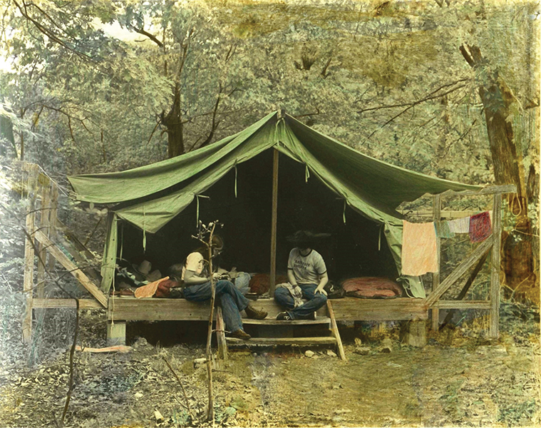This mid-1970s photo is of a green canvas tent on a wooden platform, surrounded by trees. Two counselors are perched on the steps of the platform talking with each other.