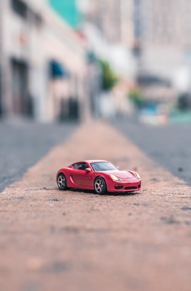 A small red toy sports car parked on a smooth, paved surface. The background is blurred, suggesting an urban setting with buildings and trees, creating a shallow depth of field effect.