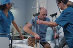 Nursing assistants help a patient get out of a hospital bed with crutches.