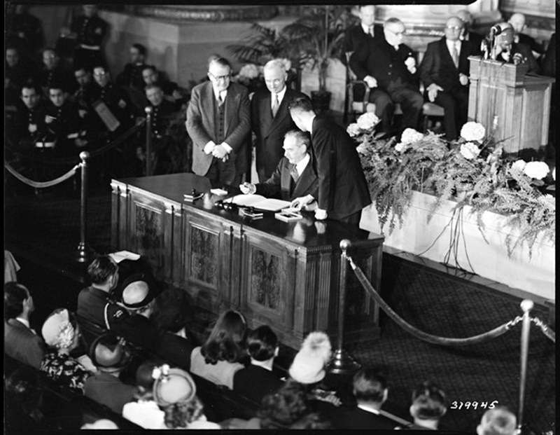 Black and white photo of people standing at a table in front of an audience, signing a document