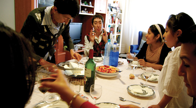 Multi-generational group of Bolivians living or visiting Pamplona, Spain are gathered around a dining table, covered with a white tablecloth, food, wine, and drinking water, are engaged in laughter and dialogue.