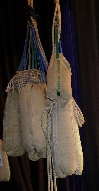 Several fabric theatrical sandbags hang by ropes backstage, used as counterweights in a hemp rigging system.