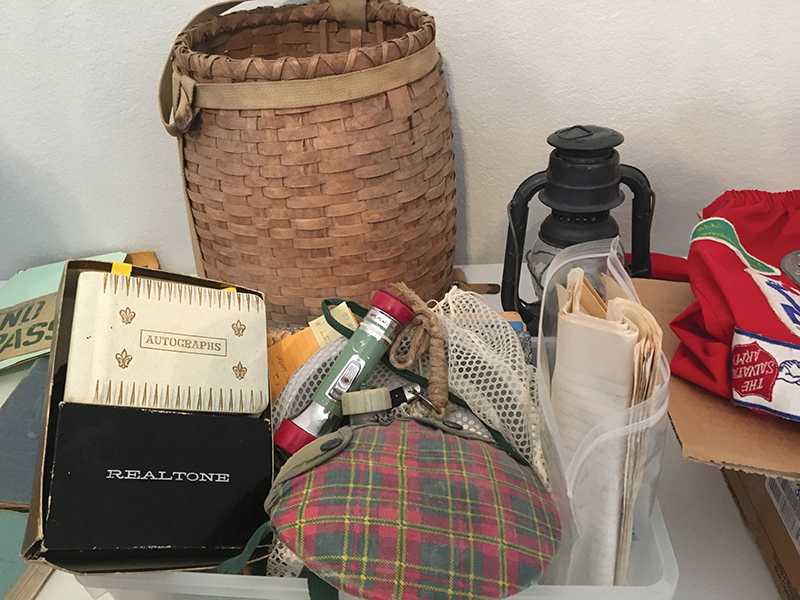 A table with various camp equipment and memorabilia, including a Girl Scout patch jacket, canteen, flashlight, wicker backpack basket, photo albums, autograph books, and a lantern.