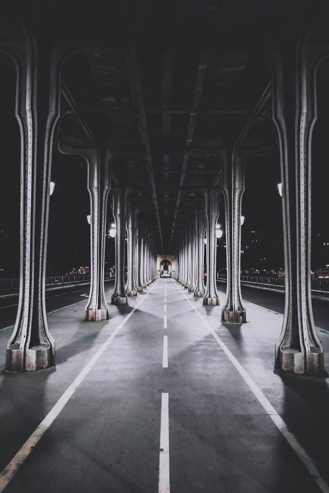 A dimly lit, deserted bridge with tall, ornate metal columns and a pathway marked by white lines. The image creates a symmetrical and tunnel-like effect, with city lights visible in the distance.
