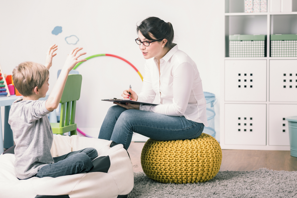A behavior analyst talks with a young child while writing notes.