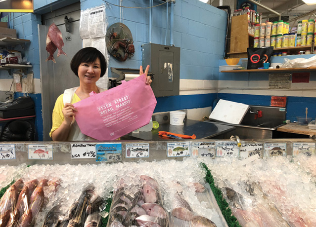 A middle-aged East Asian woman, with a short bob, stands holding a pink Essex Street Market Tote in front of a fish market stand. The woman gestures a peace sign with her left hand.