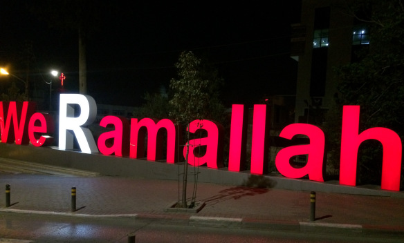Lit sign. Text: We Ramallah. The R in Ramallah is in white while the other text is red. Can be read as We R Ramallah.