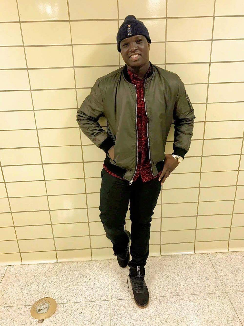 The writer, Alibamba Sillah, is shown smiling, leaning against a tiled wall and wearing a toque and leather jacket.