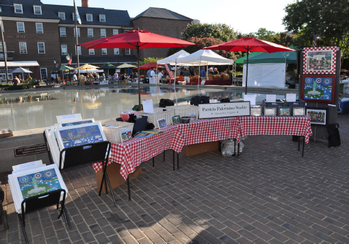 Art is displays on several tables with a sign advertising the vendor.