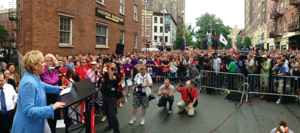 Image shows two people at a podium in front of a large crowd on a city street. One person speaks to the crowd, while the other stands next to the podium.