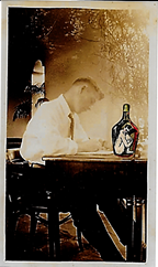 A college student sits at a writing desk on which is a bottle of whiskey which has a demon trapped inside it.