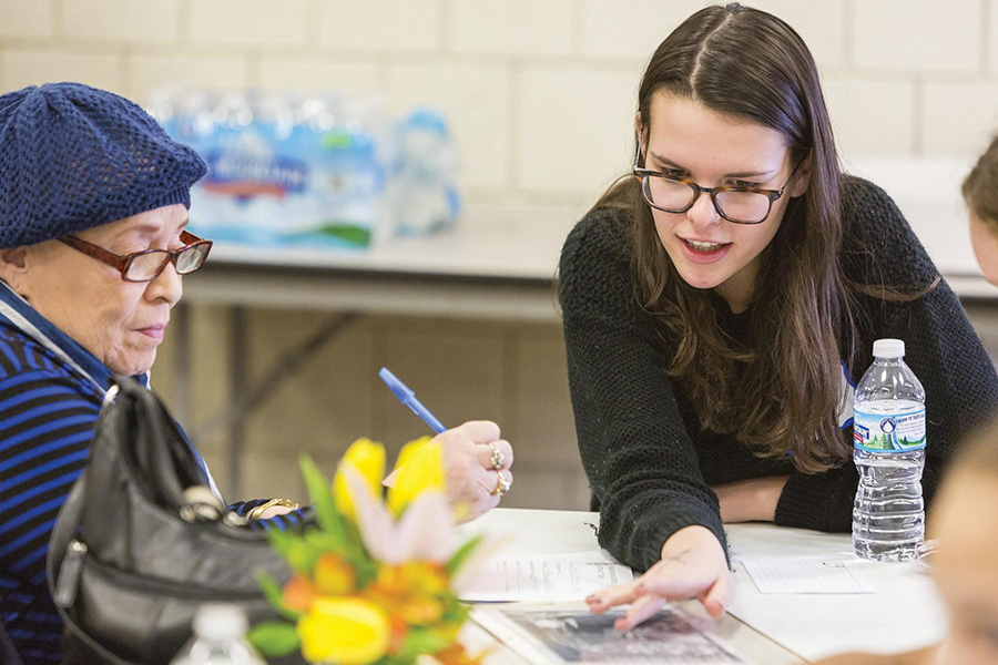 Estelle Hartshorn-Jones is dressed in a blue striped shirt and blue hat. She is holding a pen while a History Harvest student is leaning over and talking her through the process of filling out the forms.
