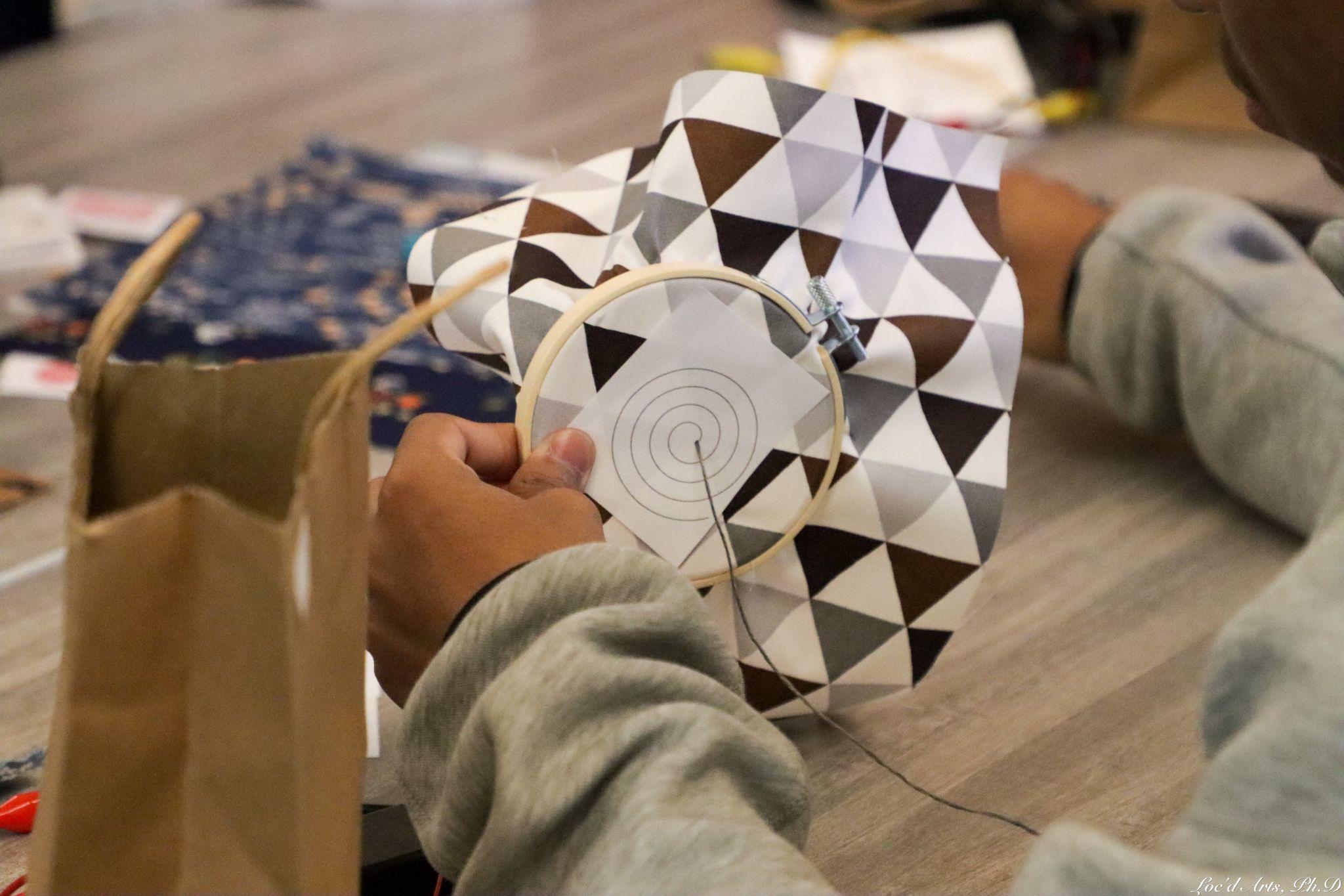 A student’s hands holding an embroidery hoop with black and white triangle-patterned fabric on it. There is a piece of paper with a spiral pattern centered in the hoop, and the student has just completed their first stitch in the center of the spiral. Other fabric is visible on the table in the background of the photo.