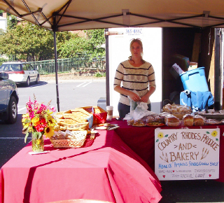 Woman stands behind her canopy stall. On it a sign reads, “Country Rhodes Produce and Bakery.”