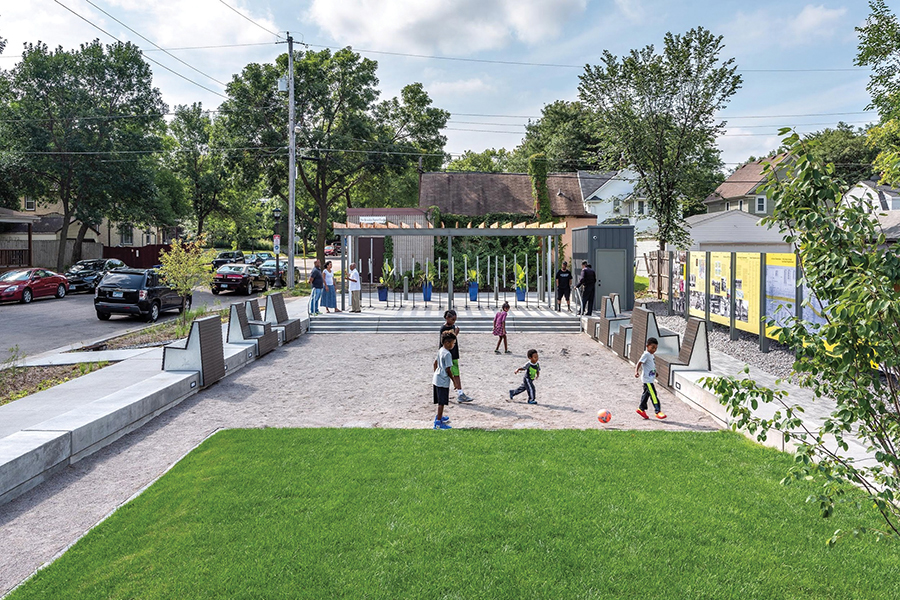 A daytime view of the commemorative plaza. In the center of the photograph, five kids are playing with a soccer ball on the dirt and grass. Along the right side, the wall of panels depicting the history stand out in bold yellow. In the background, a couple groups of adults are talking next to the art installation by Seitu Jones.