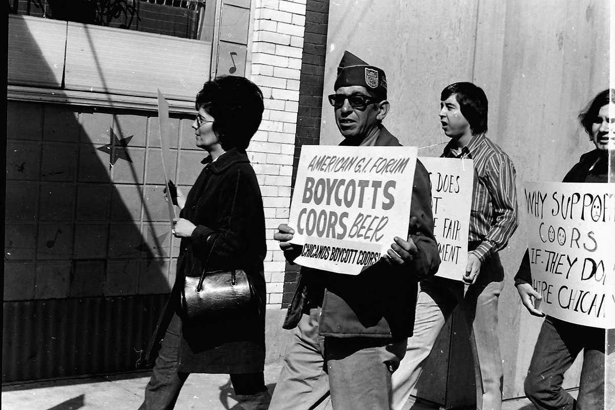 A man wearing an American GI Forum hat picketing alongside others carrying signs calling for a boycott of Coors beer