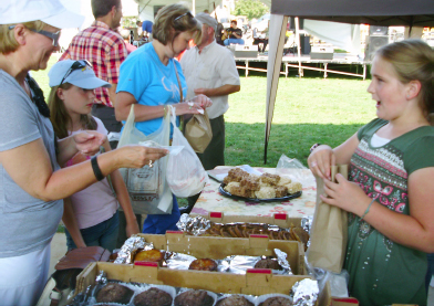 A young girl at an outside stall, happily serves a customer, putting their goods in a paper bag.