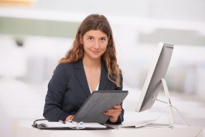 A business woman sitting at a desk with a computer monitor, an e-tablet, and a ledger.