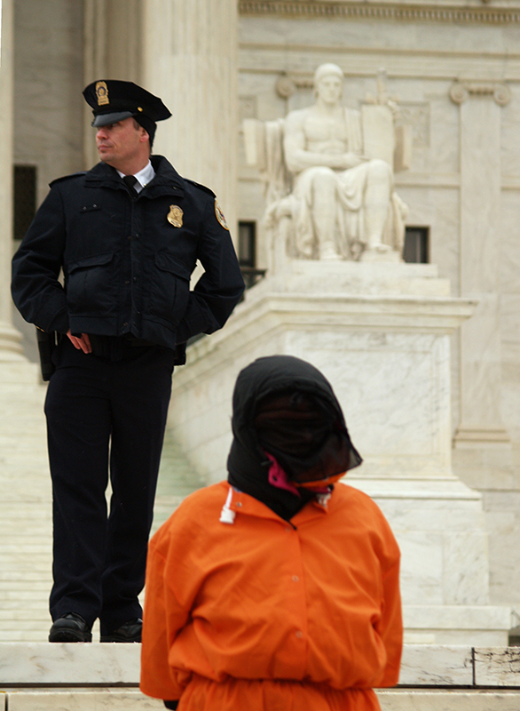Color photo of a protestor in an orange jumpsuit with a black hood over their head, in front of bored guard and a white marble statue.  