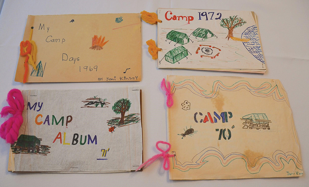 Four handmade, handwritten Girl Scout diaries, made by Joni Kinsey. The top left one is yellow with orange ribbon and has a drawing of a campfire. It is titled, “My Camp Days 1969 by Joni Kinsey”; the top right diary is titled “Camp 1972” and has a drawing of tents, a campfire, a tree and a lake nearby; the bottom left diary is titled “My Camp Album ‘71” and is decorated with drawings of a ladybug, a snake, a tent and a tree; bottom right is decorated with a drawing of a tick, a lucky rock, and a tent, and it is titled, “Camp ’70.”