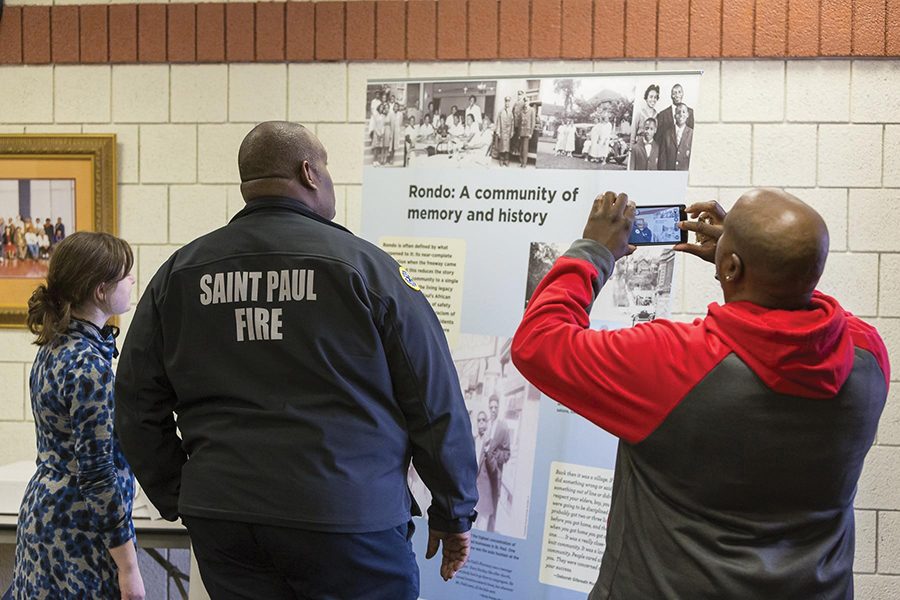 Three people face away from the camera reading a tall placard called ‘Rondo: A community of memory and history.’ Gerone Hamilton’s jacket says ‘Saint Paul Fire.