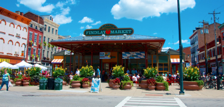 On a sunny afternoon, there are crowds of people standing outside Findlay Market front entrance. Sign on entrance reads “Historic Findlay Market”.