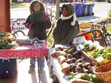 Two young girls sell produce at their stall and wave to guests.