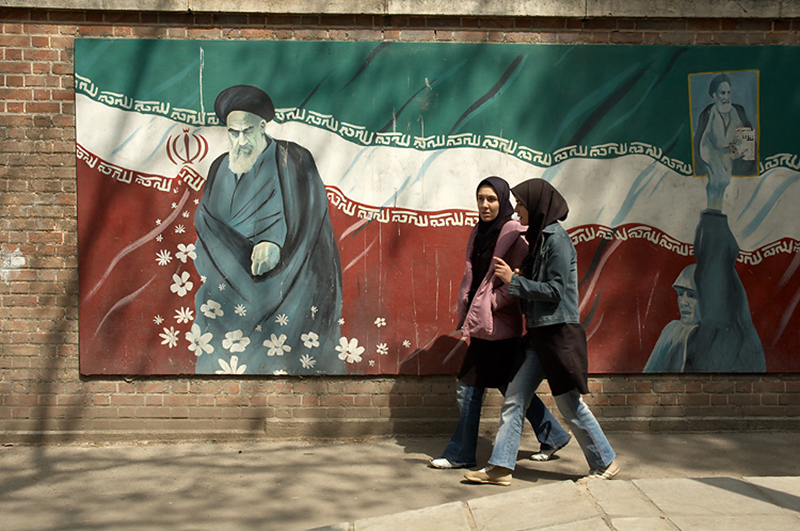 Color photo of two women walking on the street past a mural depicting Ayatollah Khomeini 