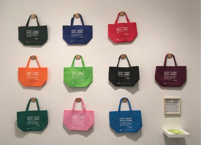Ten multicolored Essex Street Market Totes are showcased, hanging on a white wall, with a description off the right side.