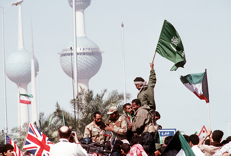 Color photo of soldiers and civilians raise American, British, Saudi, and Kuwaiti flags to celebrate following the retreat of Iraqi forces from Kuwait.