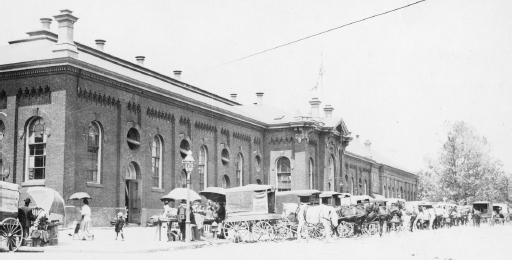 View from 7th Street circa 1914. Many carriages/buggies are parked in front of a large building.