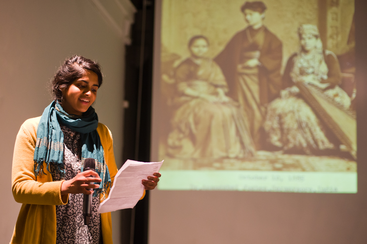 In this photo a young South Asian woman named Pavi Jaisankar, wears a turquoise scarf. She is pictured with a microphone in hand, giving a presentation at the SAADA Community Forum. An antique photo of a South Asian family of three is projected behind the young woman speaking. SAADA Community Forum, Philadelphia, PA. Taken by Vivek G. Bharathan April 4, 2013