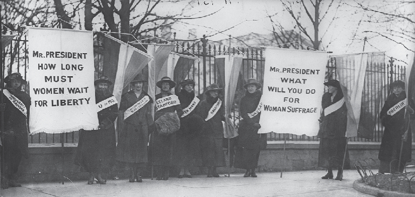 An image of a group of people marching down a street. Several pairs of people are carrying large signs between them. On both sides of the street is a crowd of observers.