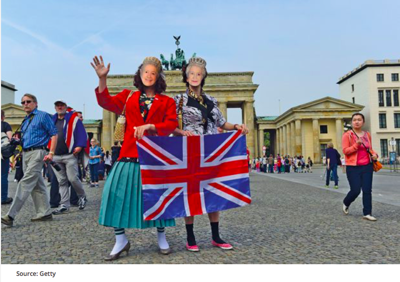 A pair of individuals dressed up and and wearing Queen Elizabeth II masks hold a union jack in front of Brandenburg Gate