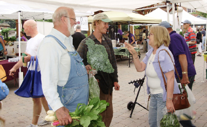 Market goers standing underneath pop up tents, holding produce and talking. A man in overalls holding up radishes and kale.