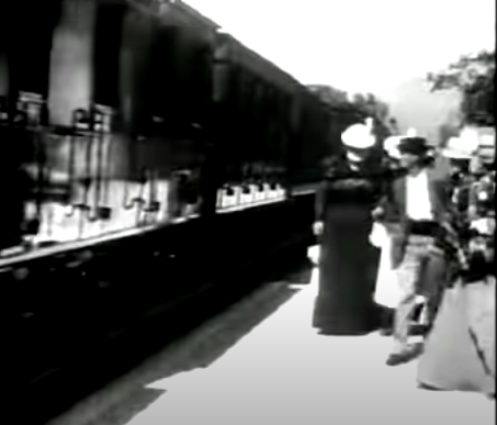 A still from a movie film showing a train on the railway tracks and two people waiting.

