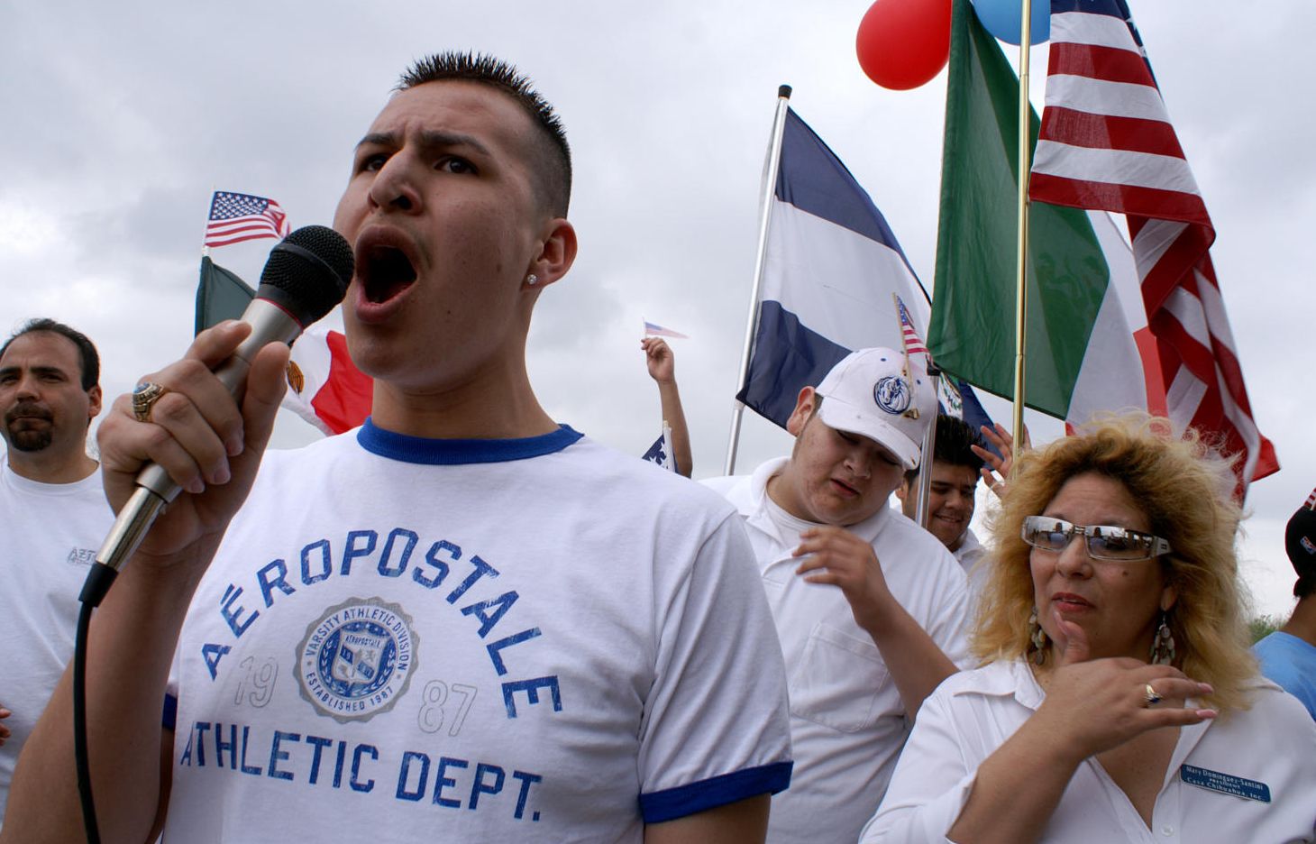 A protester speaks into microphone, while others sand behind, with flags and signs