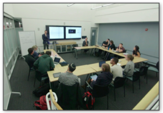 A picture of the Futurity classroom, taken by one of the authors (Hartman). Eleven students are seated around tables arrayed in a horseshoe fashion. They face a double screen at the front of the room. One screen has a PowerPoint projected onto it and one screen has Lacuna projected on to it. The instructors flank the screens, one sitting and one standing. Four students have laptops open, but all appear to be engaged in the conversation.