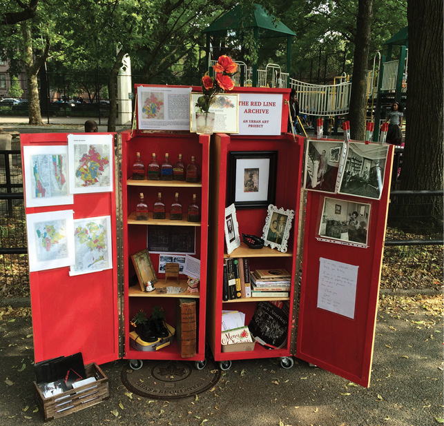 Pictured here is artist Walis Johnson’s Red Line Archive. A mobile cabinet is displayed in a park; the cabinet is painted and appears with open doors; inside are objects like maps, framed photos, shoes, books and liquors that represent the pre-gentrified Black population of Brooklyn, NY. Atop the cabinet are flowers setting in a cup.