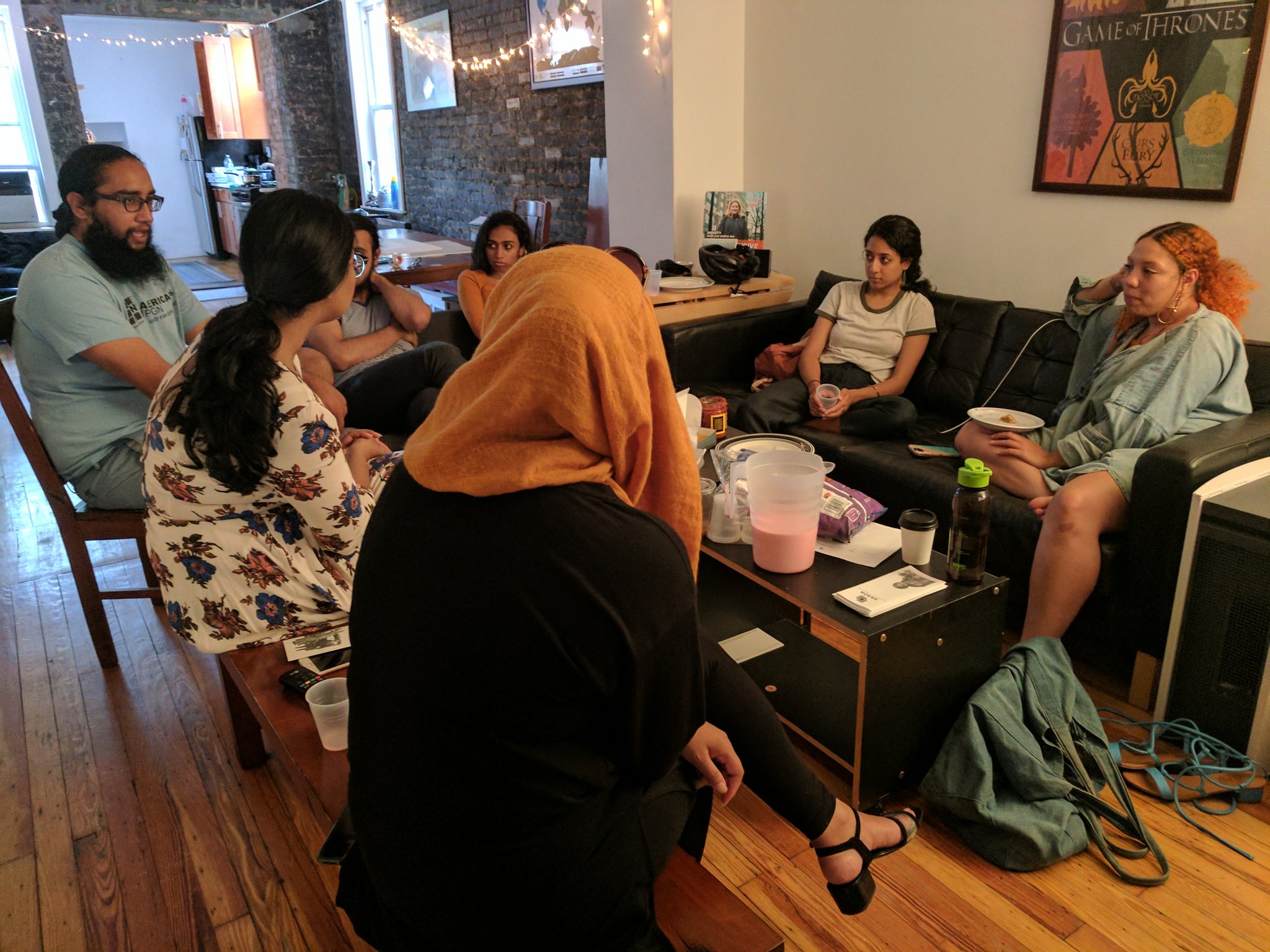Pictured here are seven South Asian young adults sitting in a community member’s living room, gathered around a coffee table and seated on a black sofa, engaging in dialogue