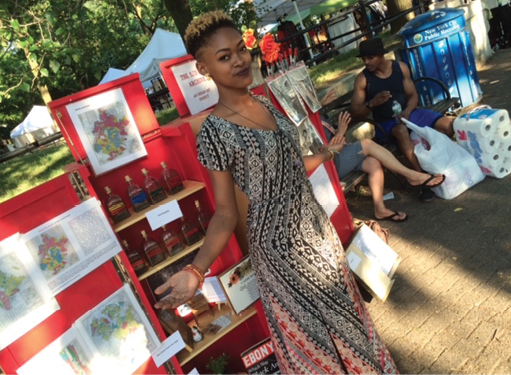 This is a photo of a young Black woman with short-cropped natural hair. This woman is wearing a long dress and she is pictured modeling in front of the Red Line Archive.